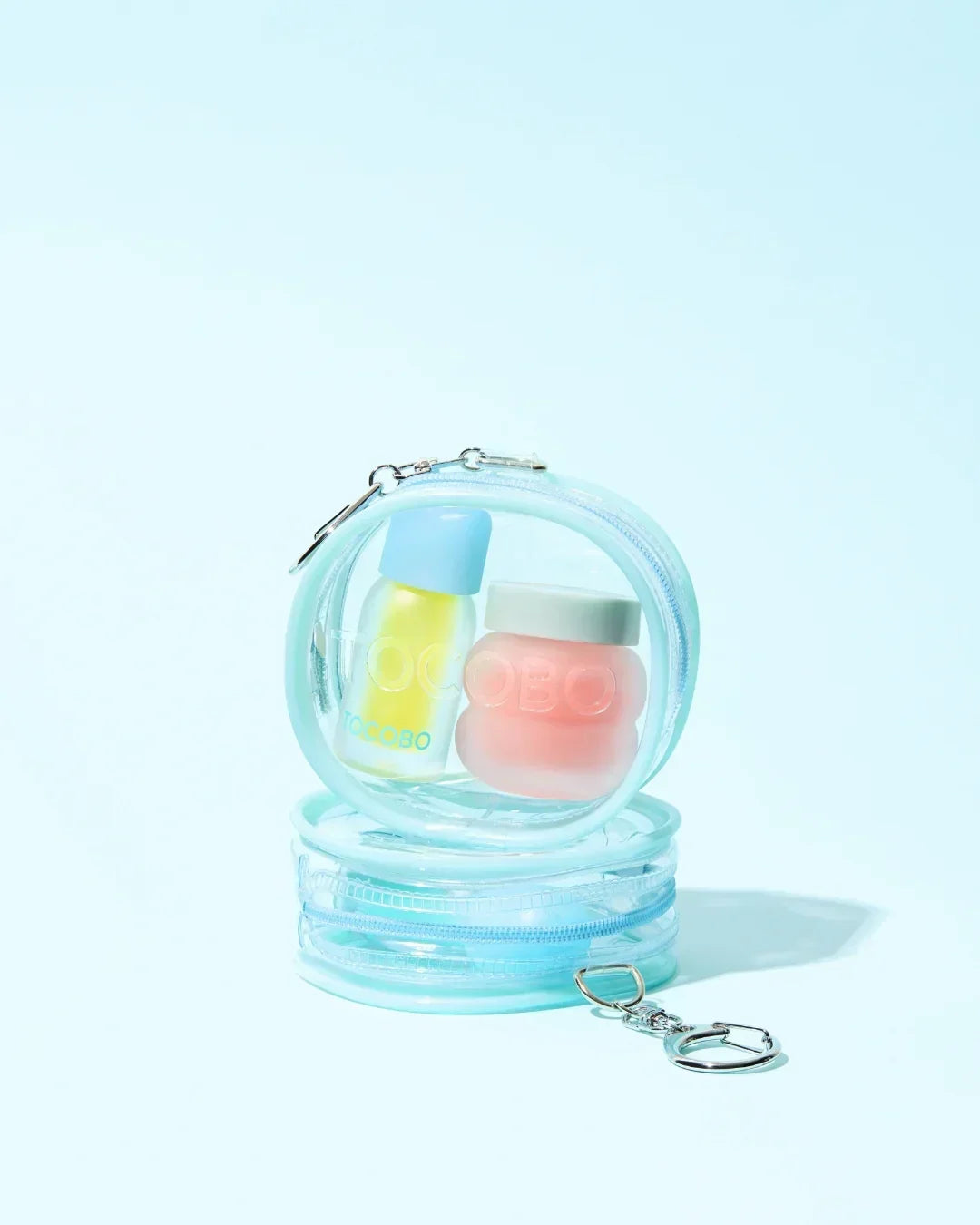 Transparent blue pouch keyring holding a yellow lip oil and a pink lip balm container on light blue background