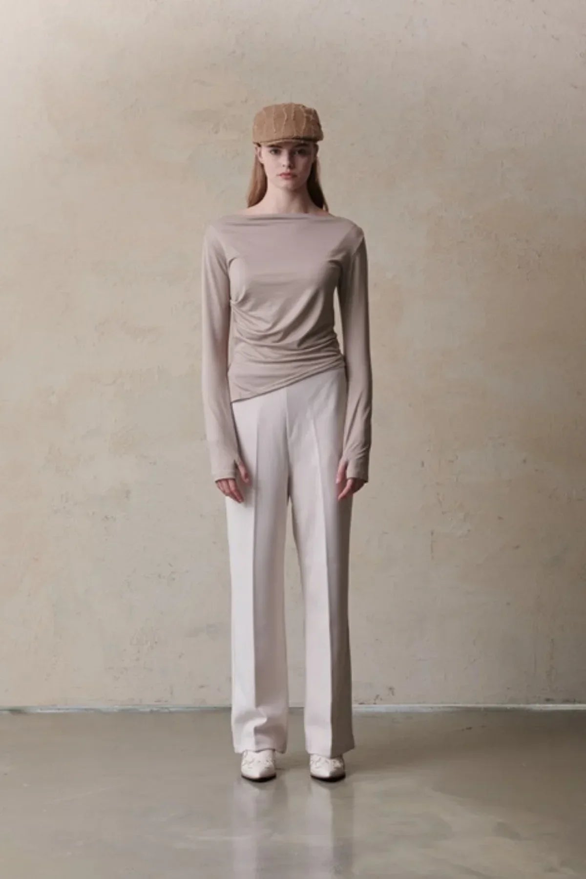 Woman wearing beige long-sleeve top, light cream wide-leg pants, white shoes, and brown textured cap in a minimalist setting