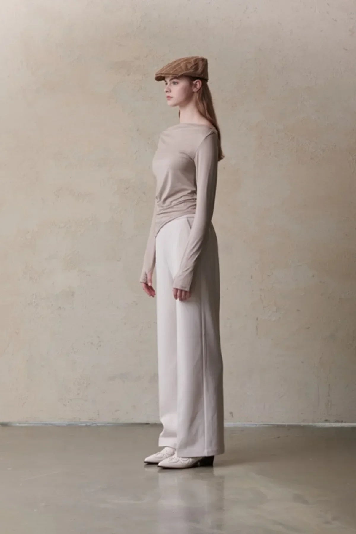 Side view of woman wearing beige long-sleeve top, white wide-leg pants, tan cap, and white shoes against textured wall