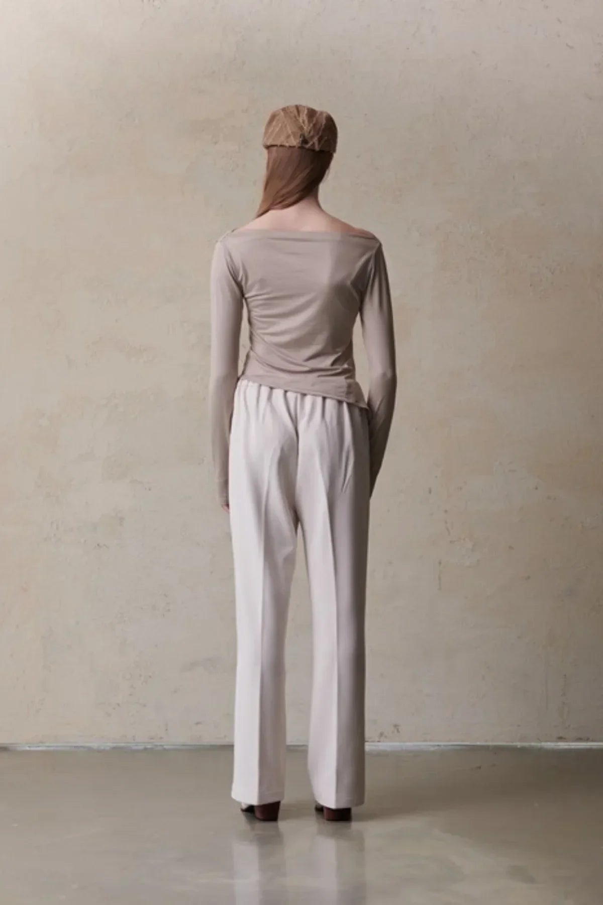 Woman in beige long-sleeve top, white wide-leg pants, and brown beret standing against neutral wall