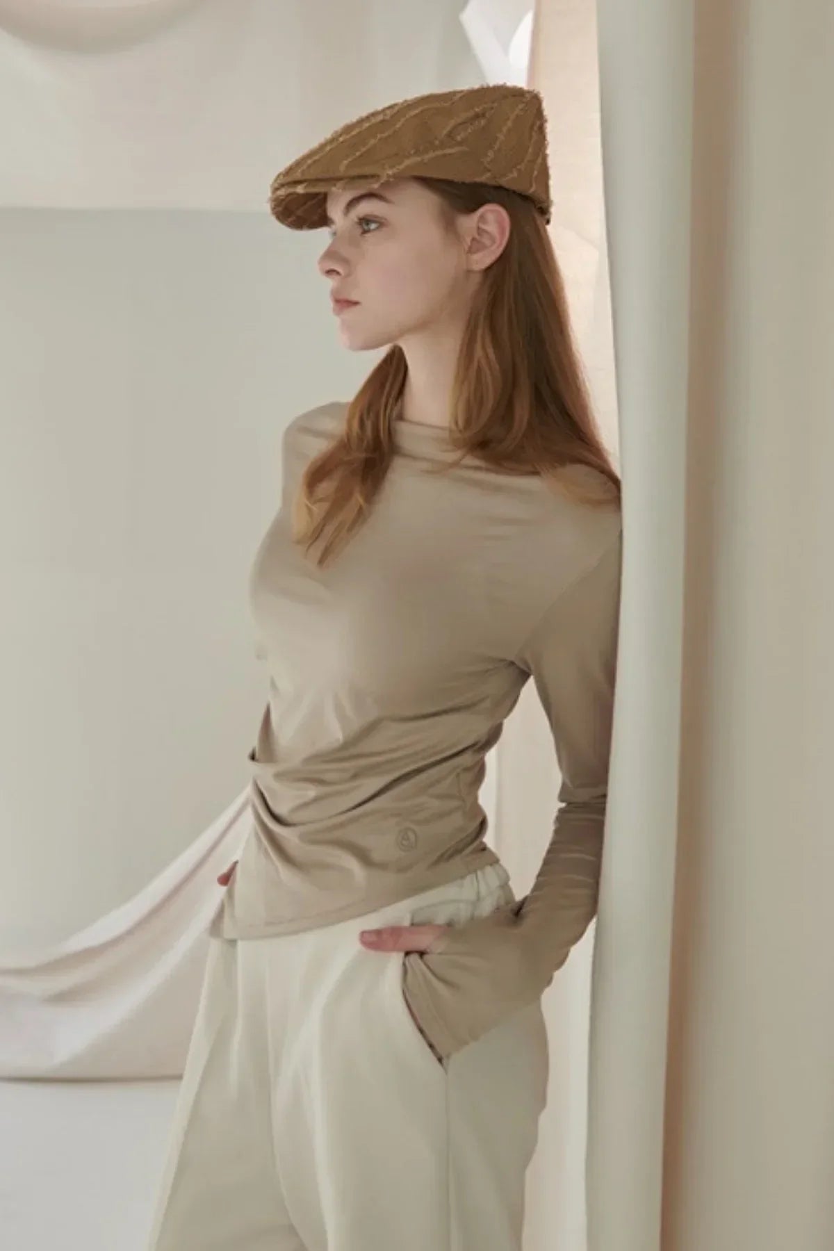 Woman wearing beige long-sleeved top and light cream pants, standing indoors with hands in pockets