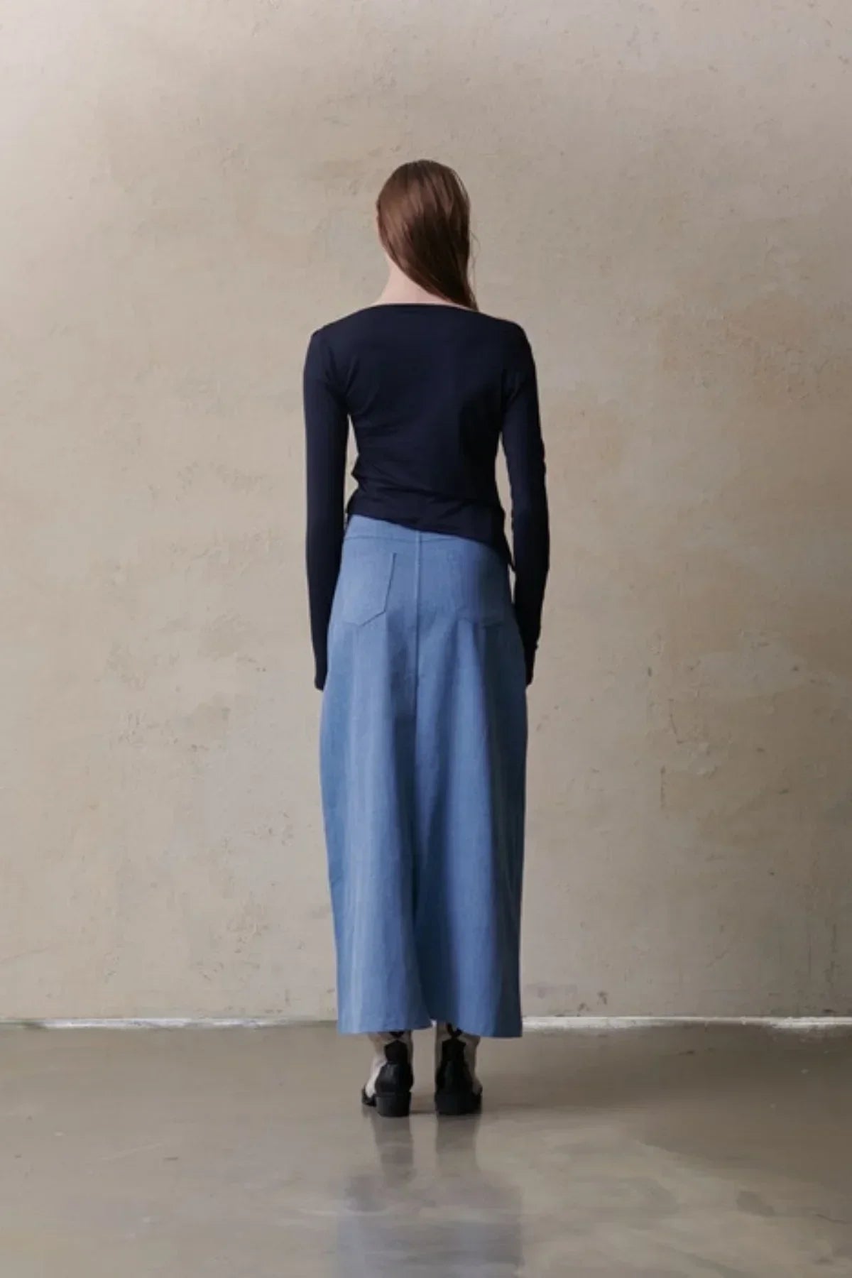 Woman in navy long-sleeve top and light blue midi skirt standing against textured wall