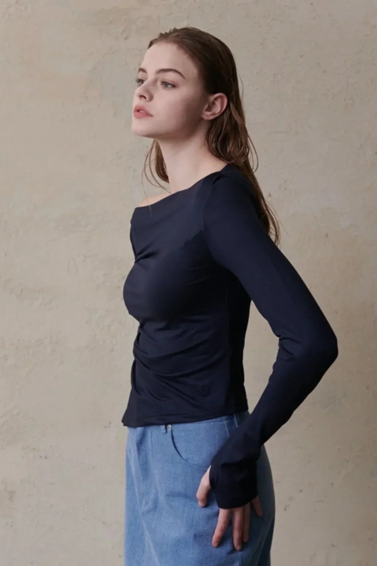 Woman wearing navy blue long-sleeved top and light blue jeans against a textured beige wall