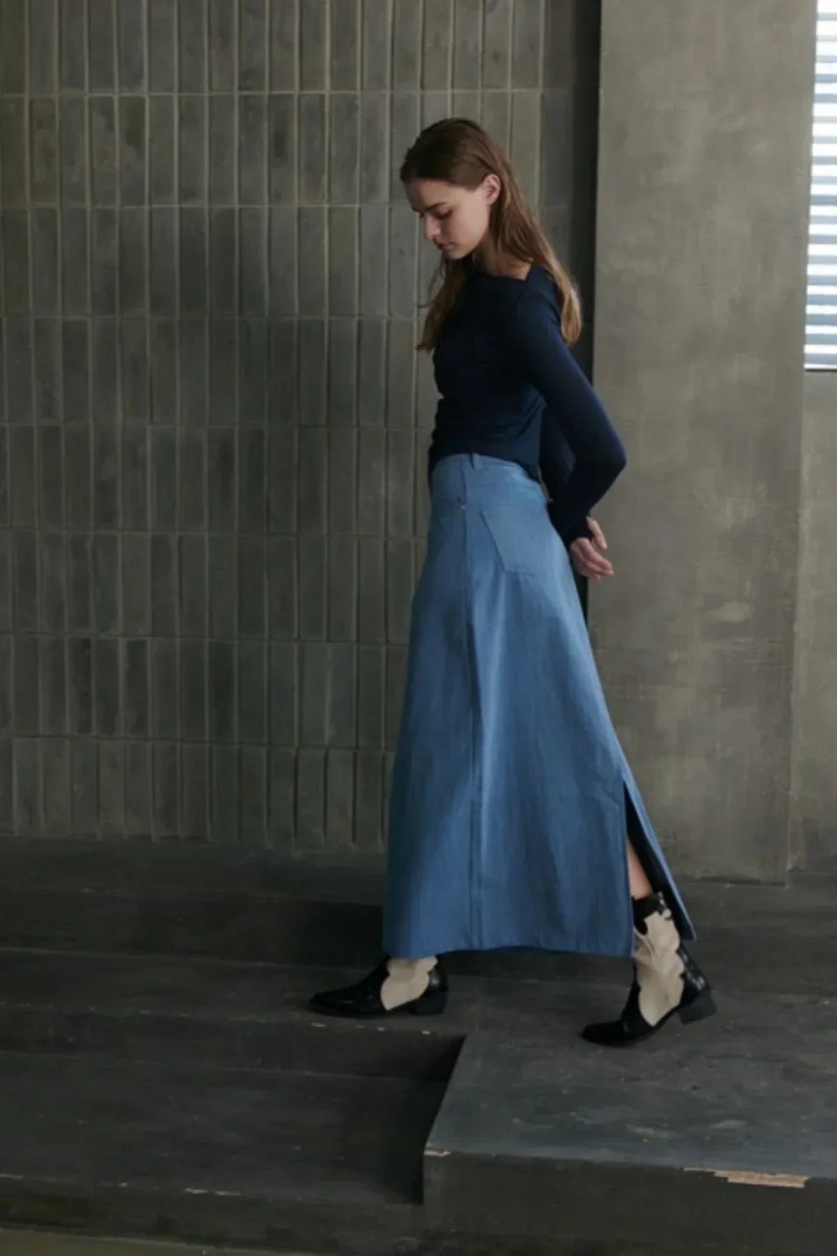 Woman in navy long-sleeve top and light blue denim maxi skirt with black and white ankle boots in industrial setting