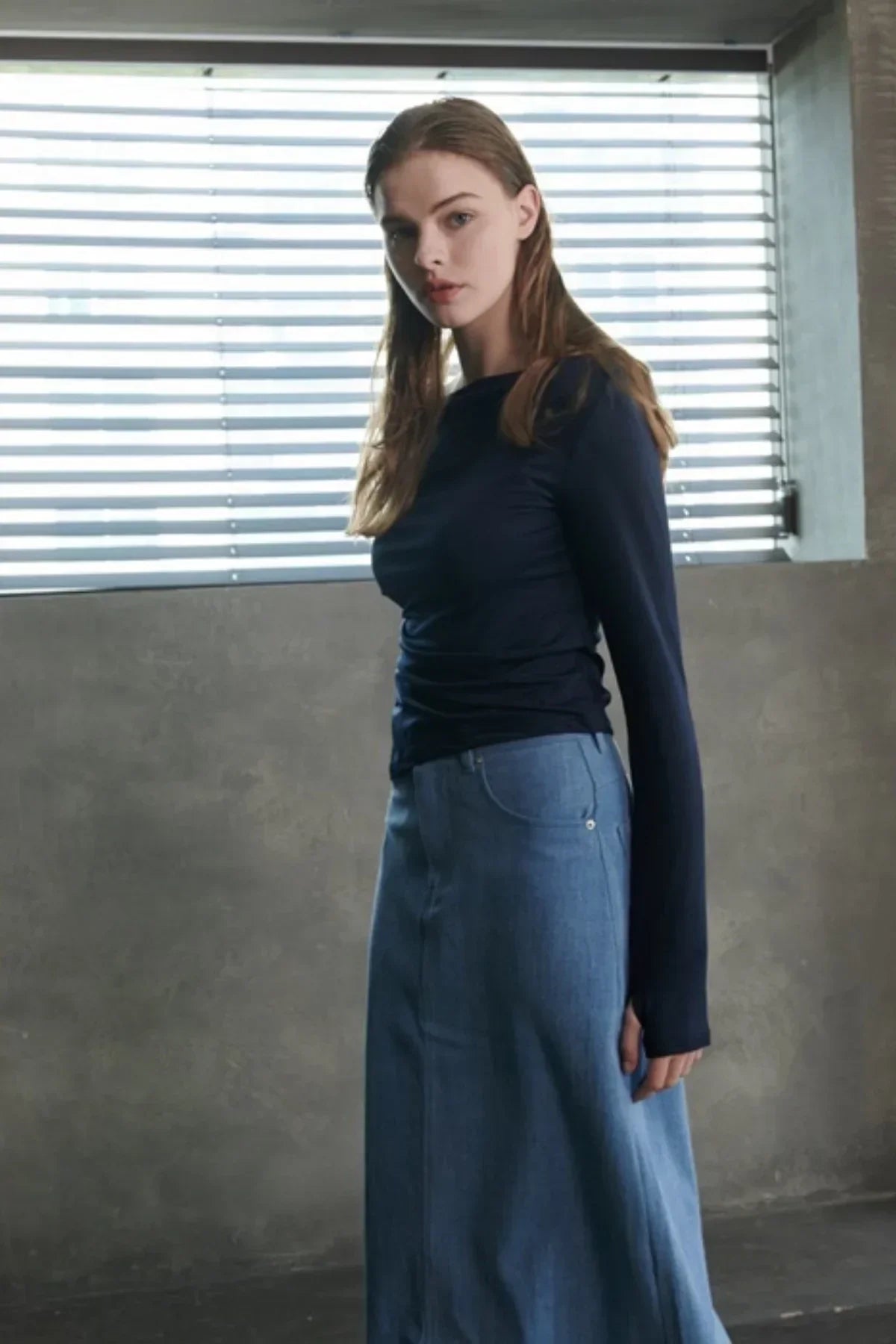 Woman wearing navy long-sleeve top and high-waisted light blue denim skirt near window blinds