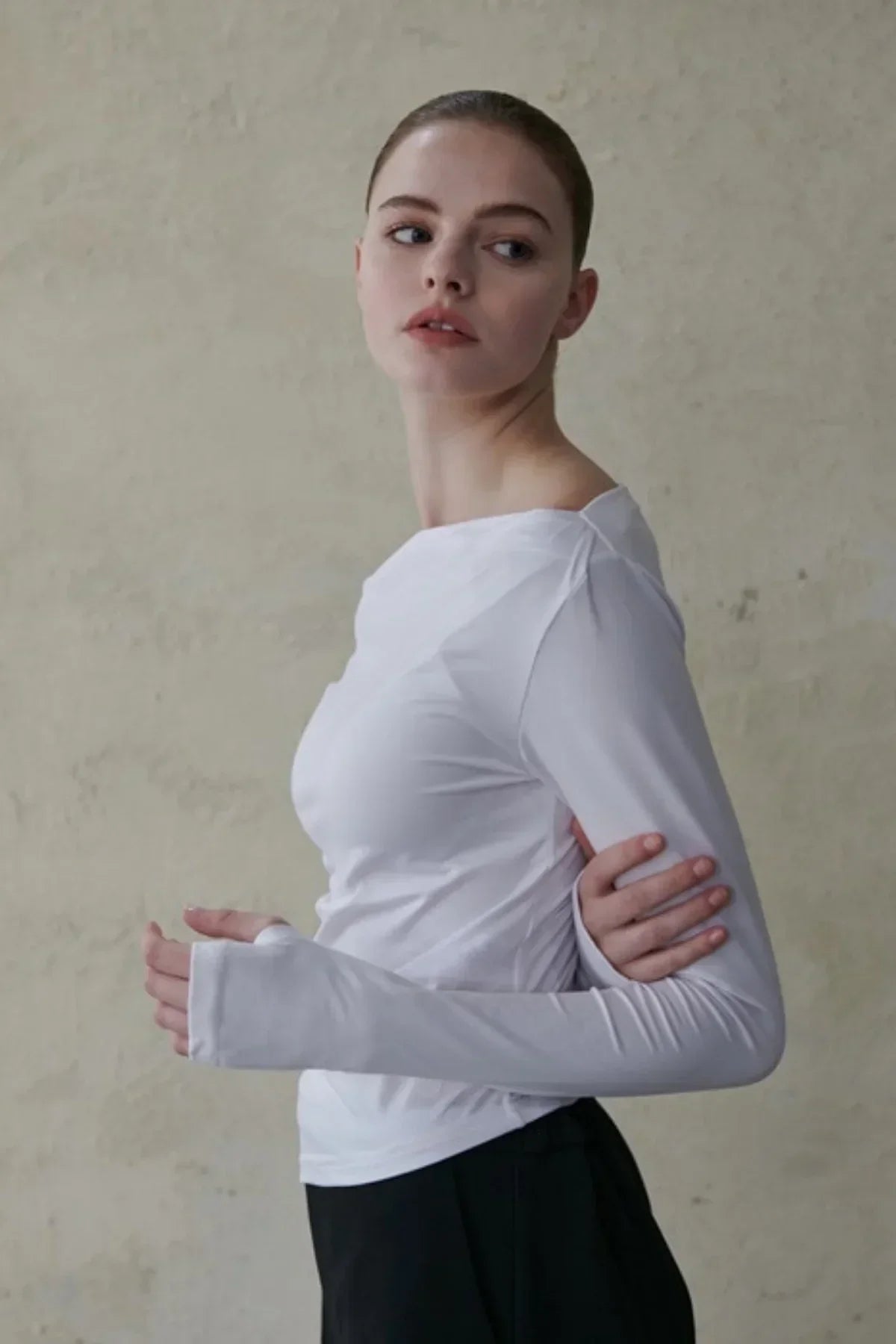Portrait of a woman wearing a fitted white long-sleeve top with thumb holes against a textured beige wall