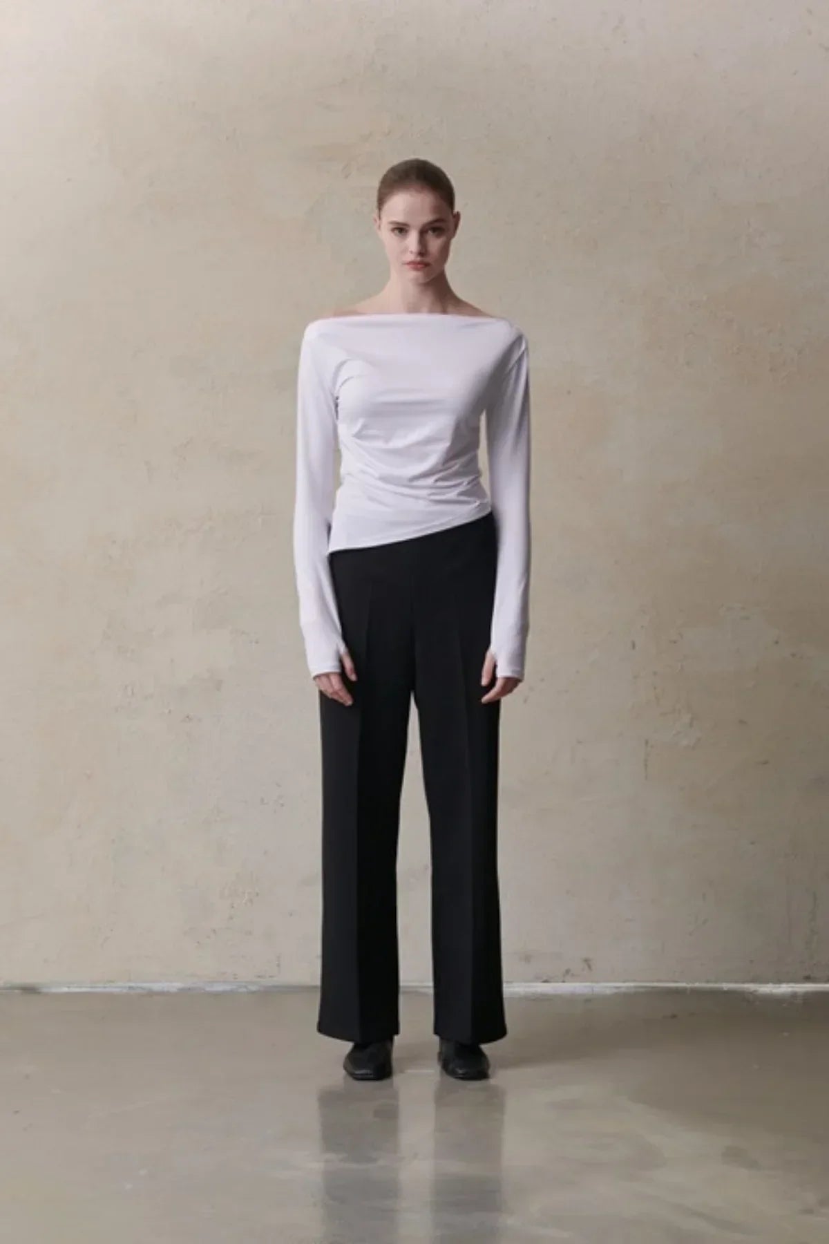 Woman wearing a white long-sleeve off-shoulder top and black wide-leg pants against beige wall