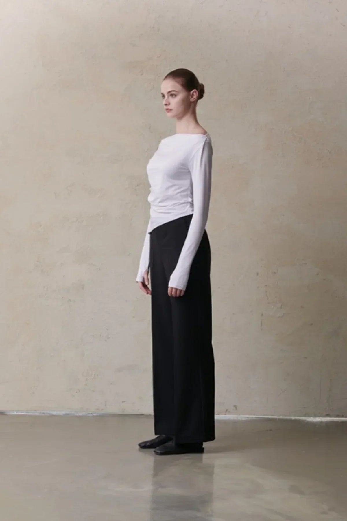 Side view of female model wearing white long-sleeve fitted top and black wide-leg pants against beige wall