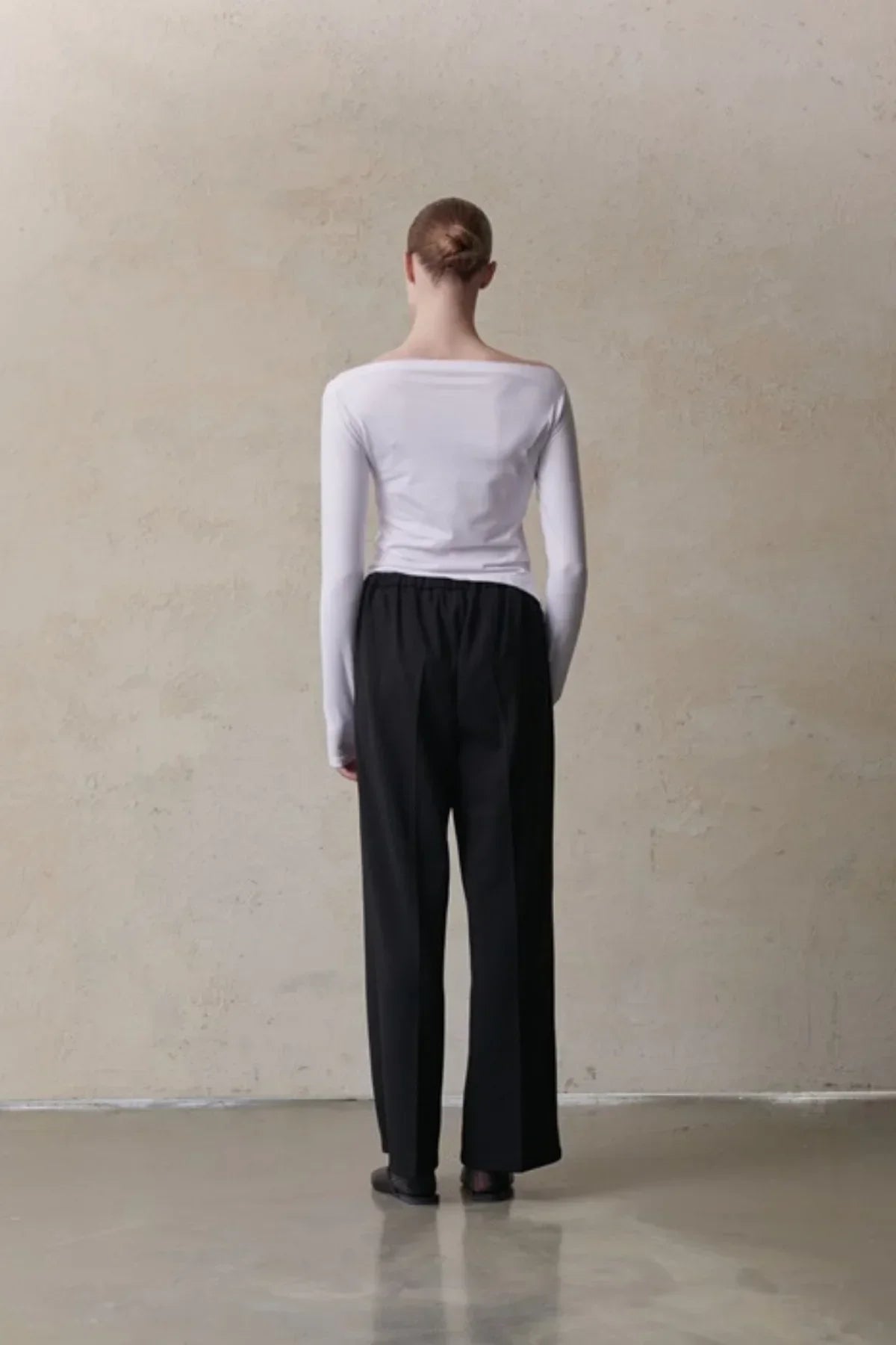 Back view of a model wearing a white long-sleeve top and loose black pants against a plain beige wall