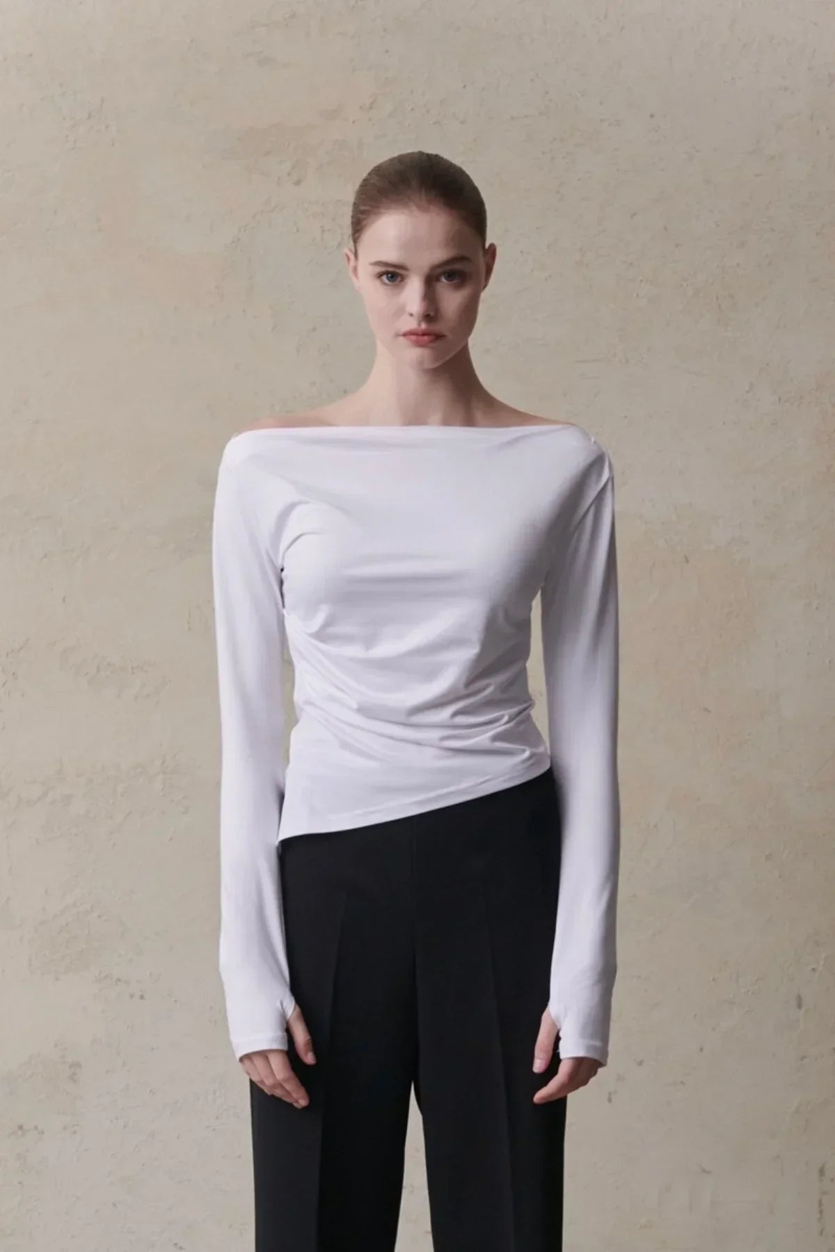 Woman in white long-sleeve off-shoulder top and black pants standing against beige textured wall