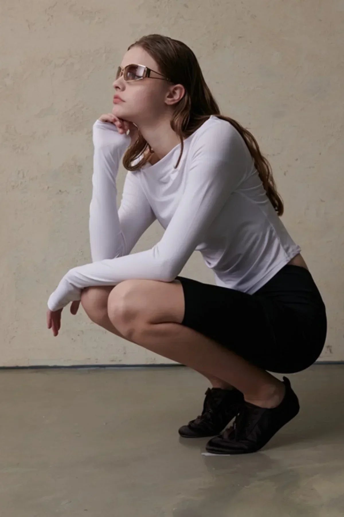 Woman wearing white long-sleeved fitted top and black shorts crouching against textured beige wall