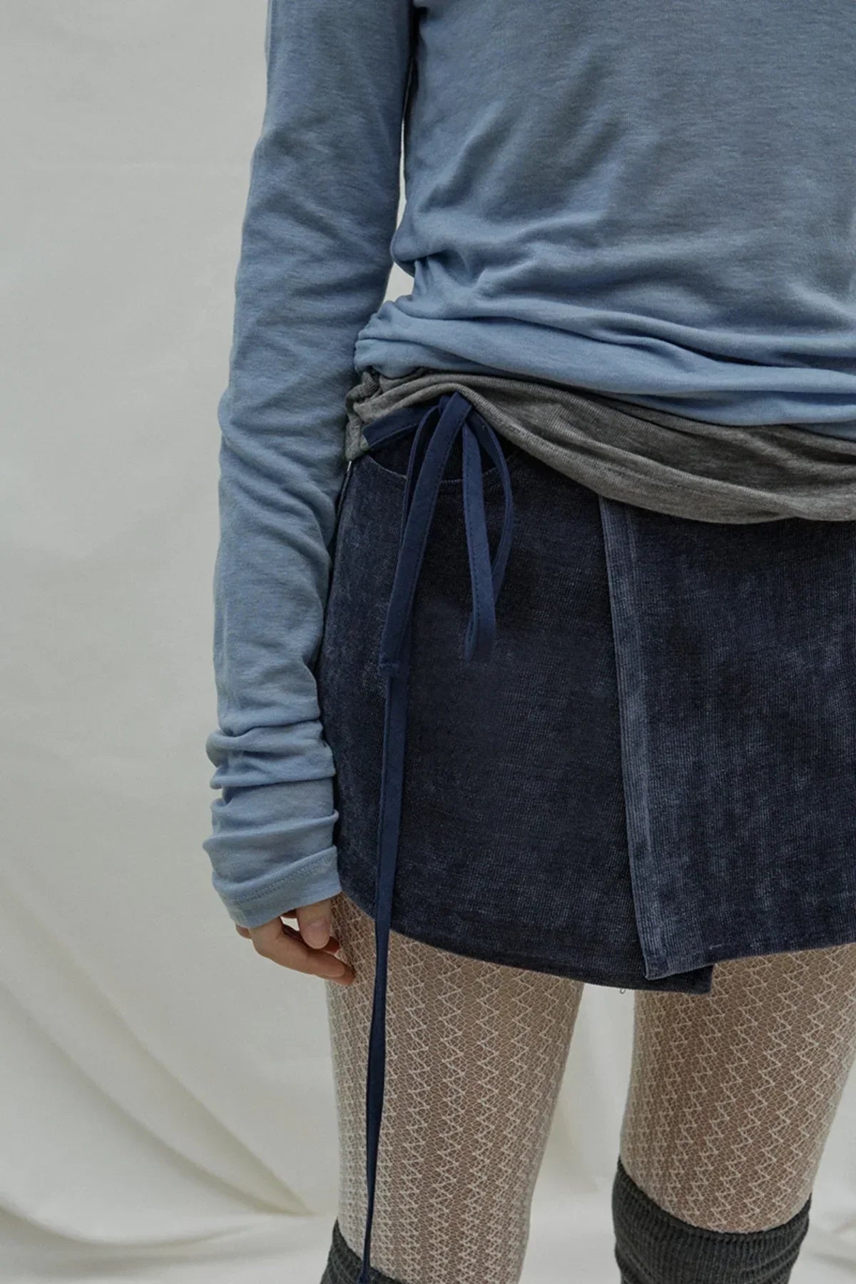 Close-up of a person wearing a cloudy blue long-sleeve top, navy wrap skirt, patterned tights, and dark leg warmers