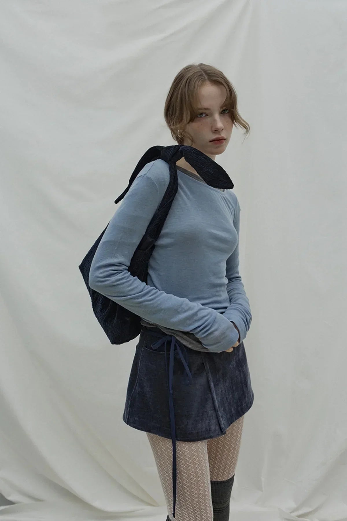 Young woman wearing cloudy blue long sleeve top, dark mini skirt, knitted tights, and black shoulder bag