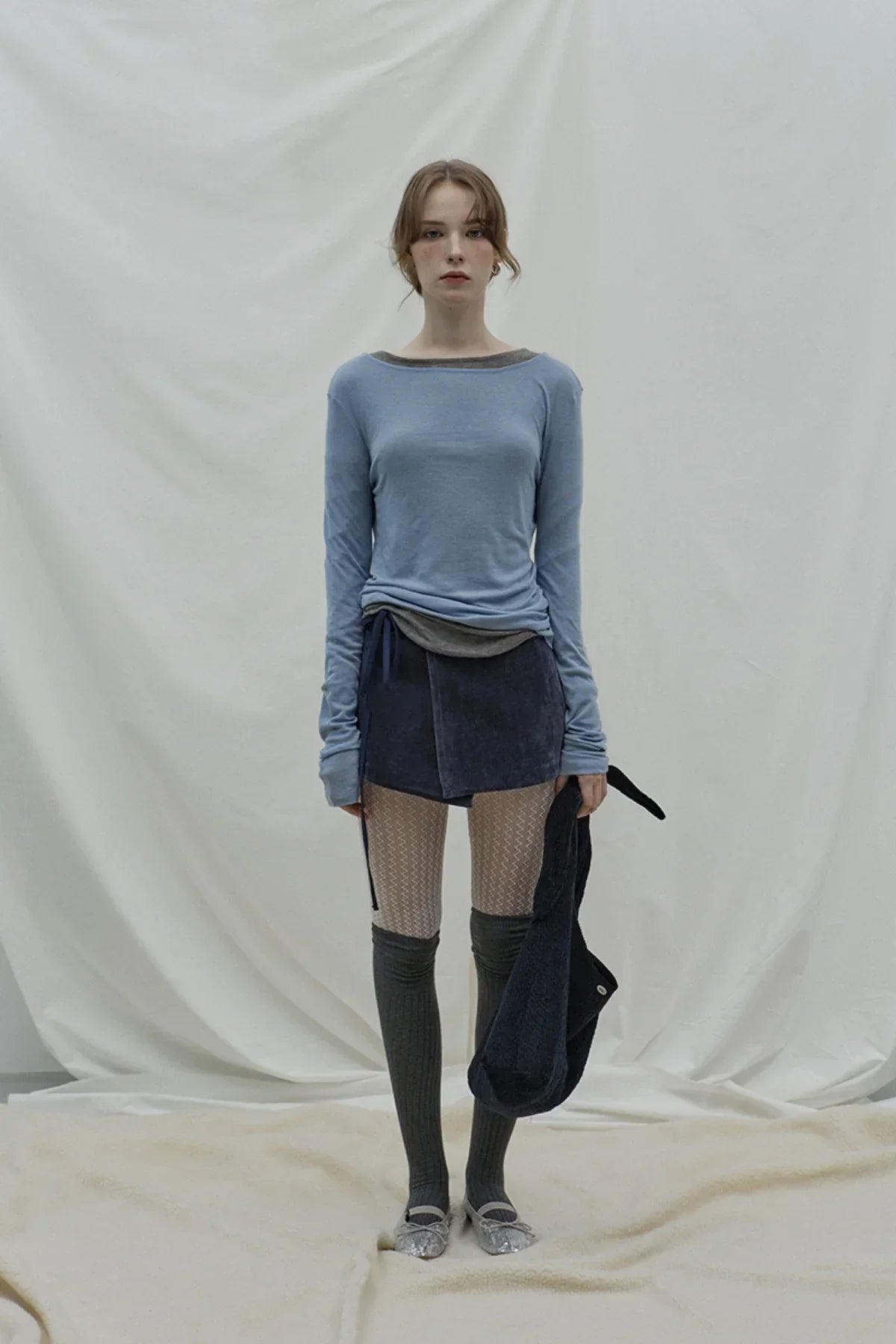 woman in cloudy blue long sleeve top, navy wrap skirt, patterned tights, knee-high socks, and silver flats on cream backdrop