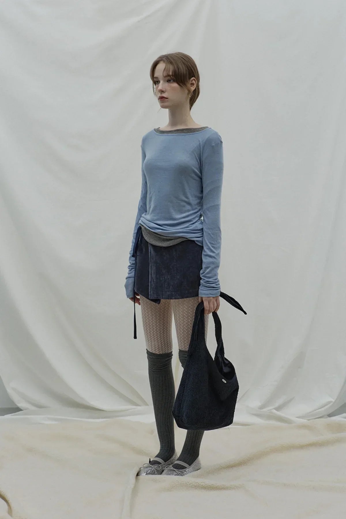 Model wearing cloudy blue long-sleeve top, dark mini skirt, patterned tights, knee-high socks, holding black bag