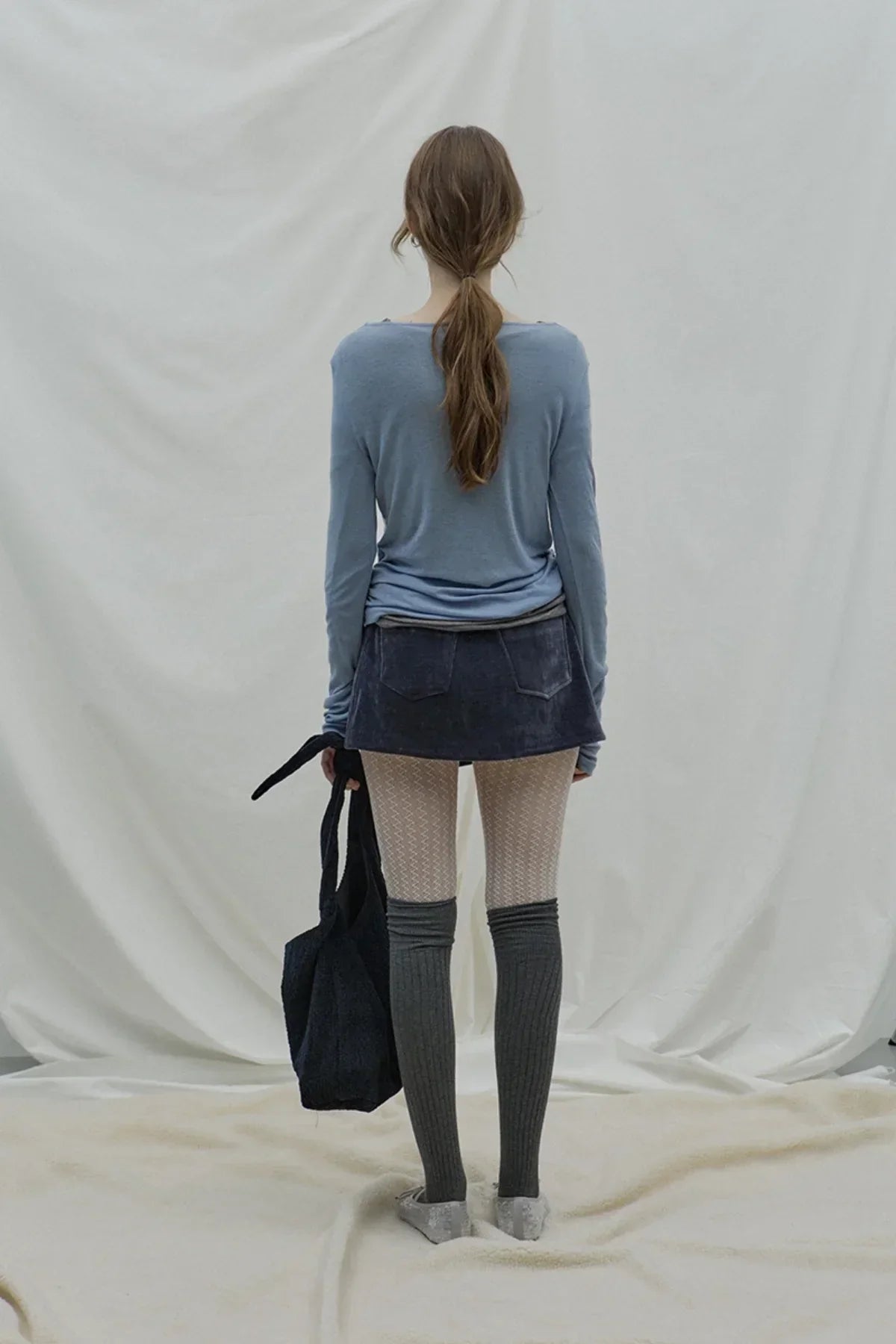 Rear view of woman wearing cloudy blue loose fit long sleeve top, navy mini skirt, patterned leggings, and dark knee-high socks, holding black tote bag