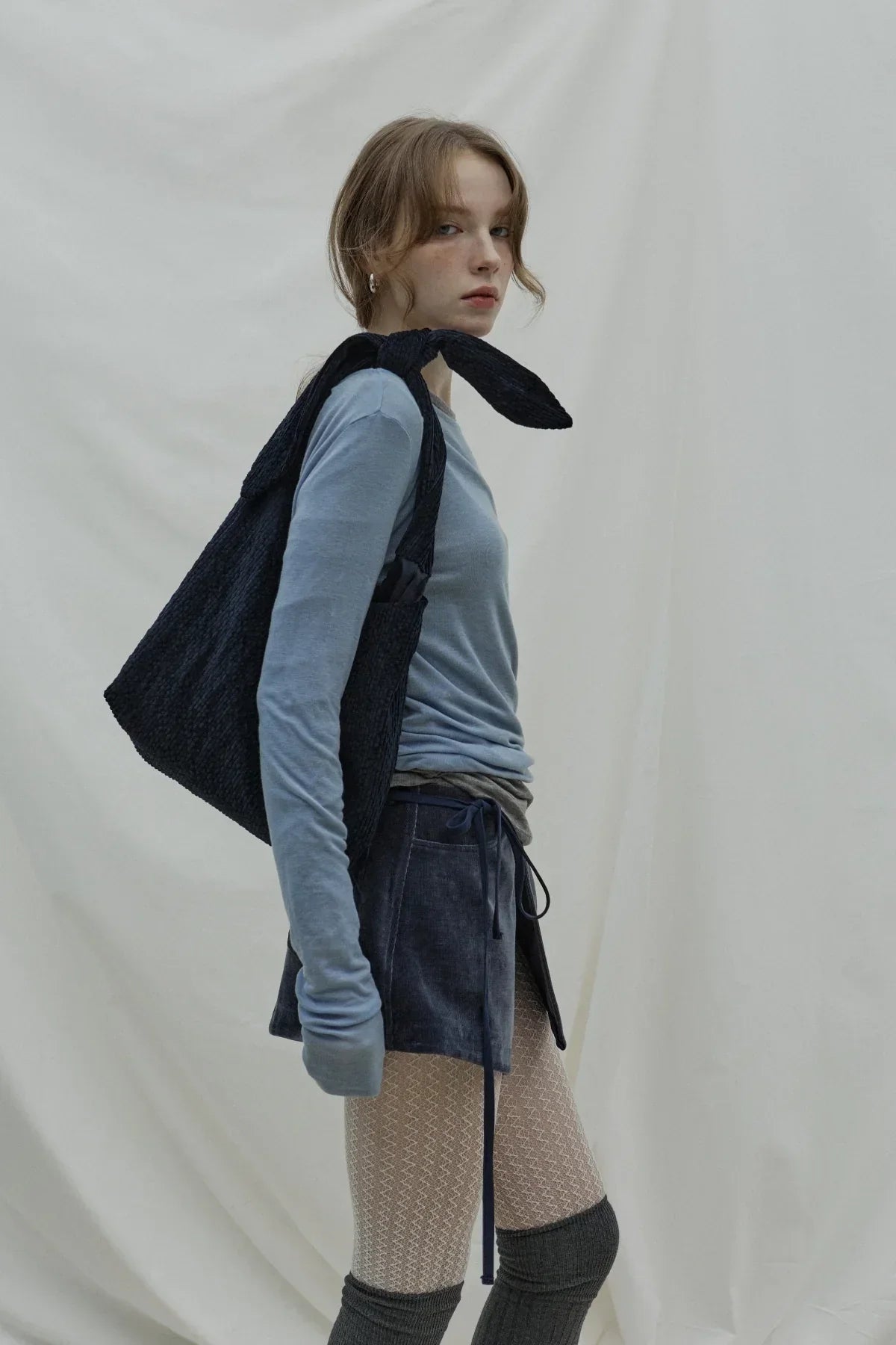 Young woman in cloudy blue long sleeve top, textured navy skirt, patterned tights, and black bag in studio