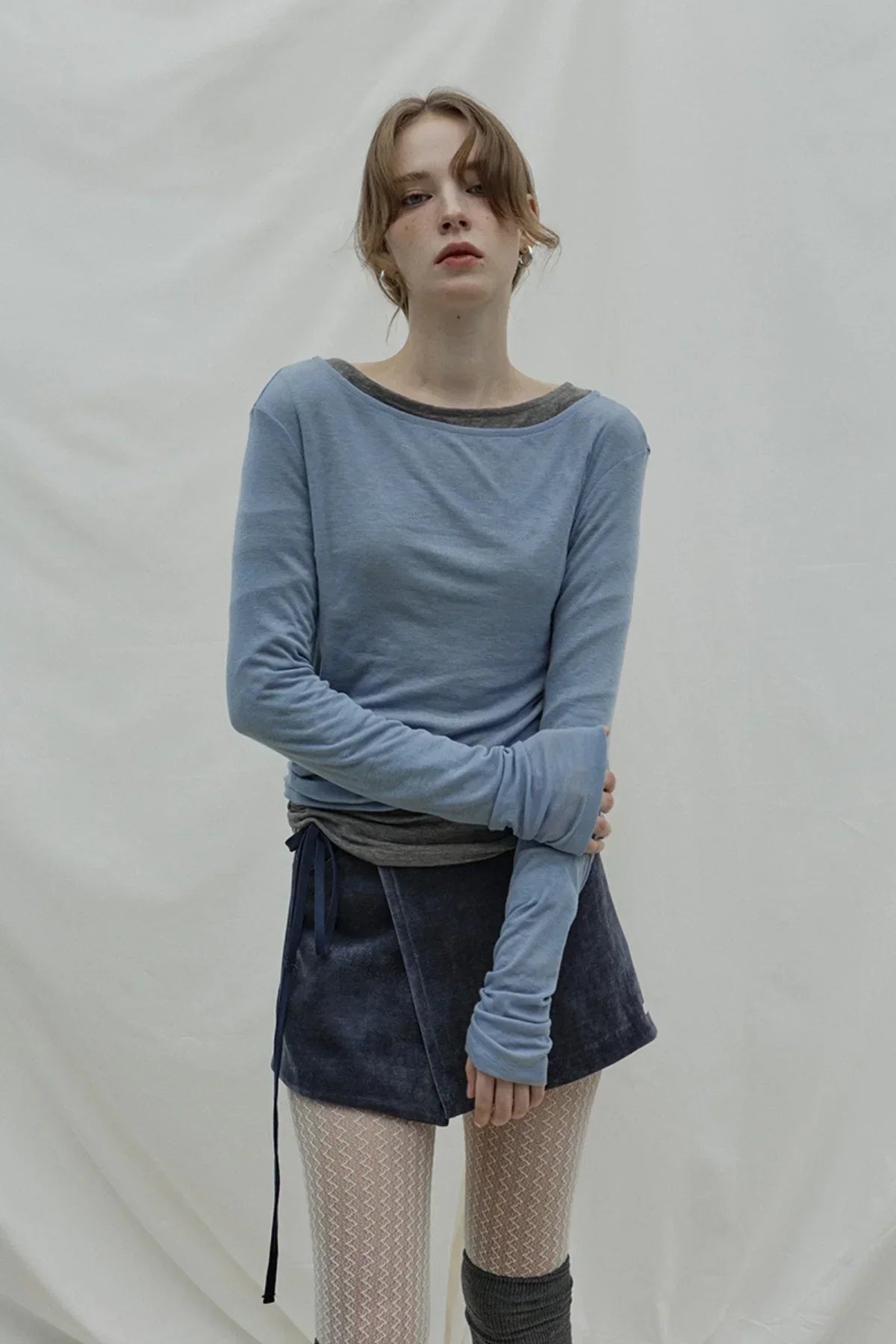 Woman in loose fit cloudy blue long sleeve top with navy skirt and patterned tights on plain background