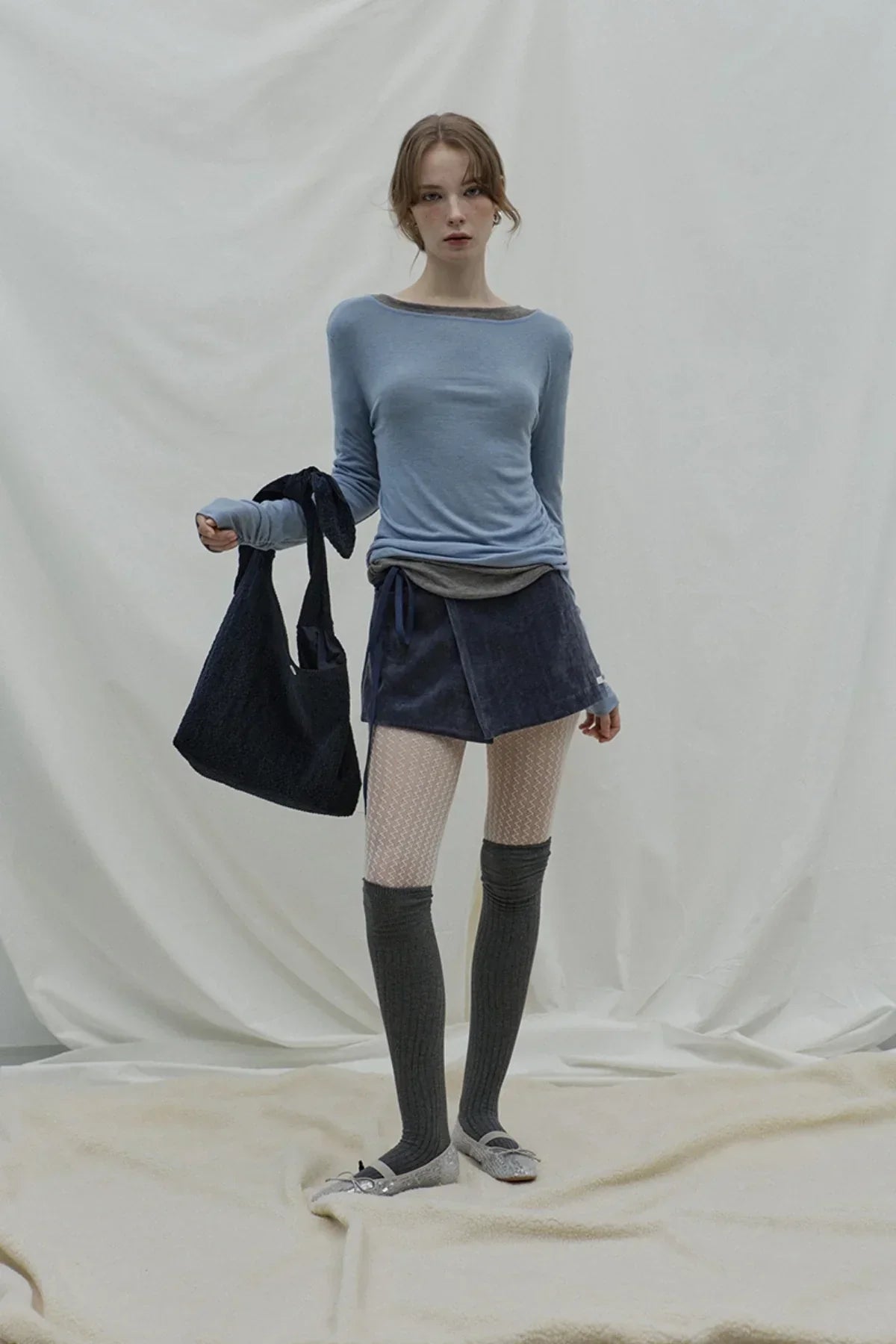Fashion model in loose fit cloudy blue long-sleeve top, navy wrap skirt, patterned tights, knee-high socks, and silver flats