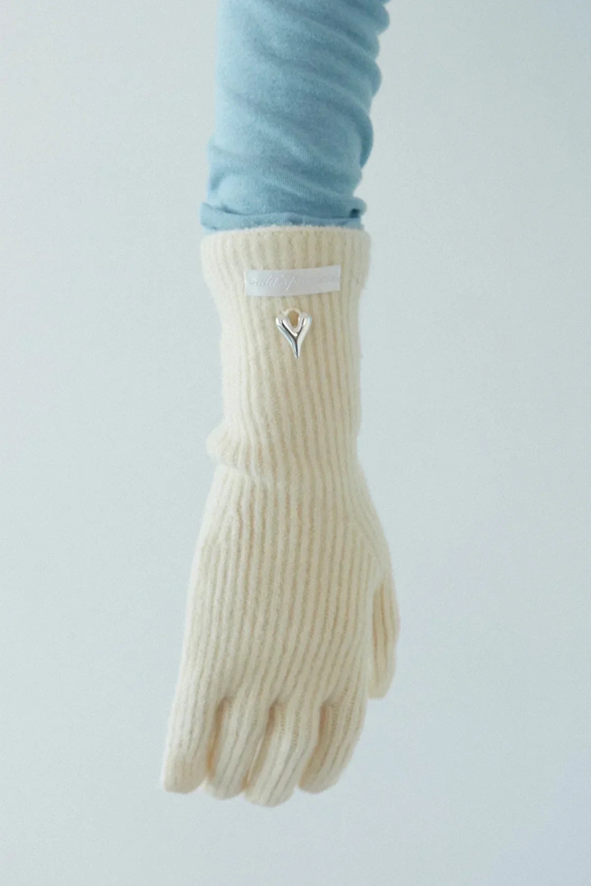 Close-up of a hand wearing cream ribbed knit gloves with a silver heart emblem and light blue sleeve