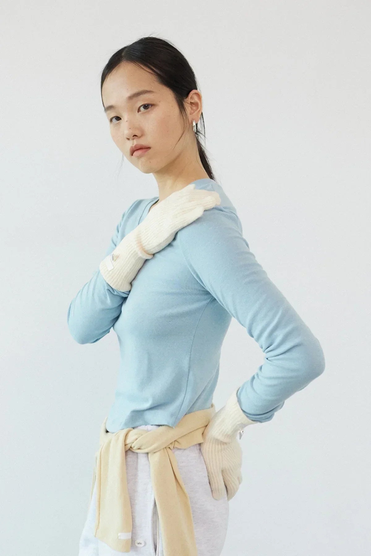 Woman wearing cream knit gloves, light blue long-sleeve top, and beige sweater tied at waist