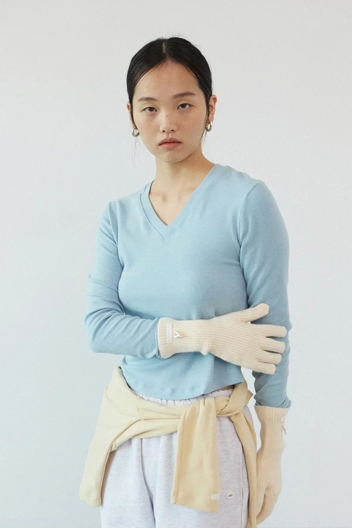 Young woman wearing cream ribbed gloves, light blue long sleeve top, and beige sweater tied around waist