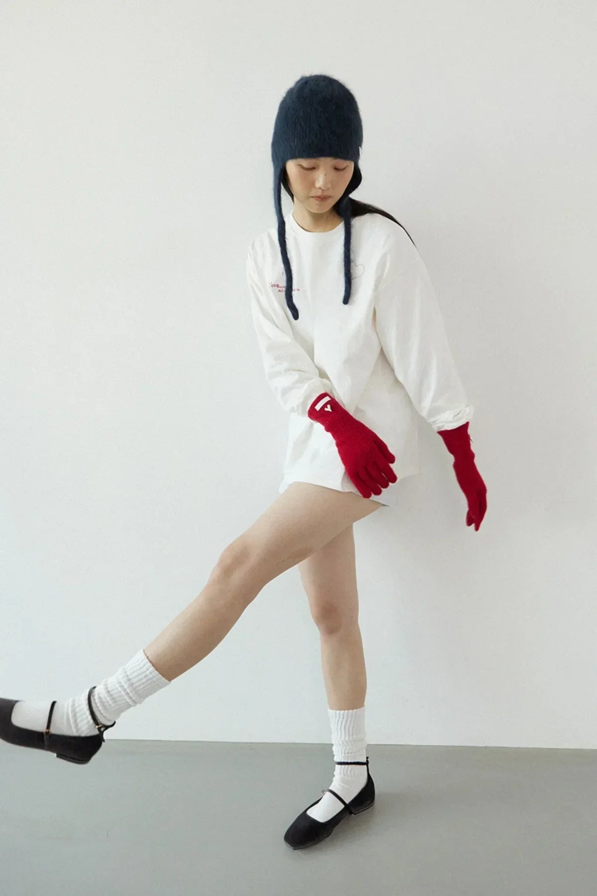 Young woman wearing red gloves, white long sleeve shirt, black Mary Jane shoes, and navy knit hat posing indoors