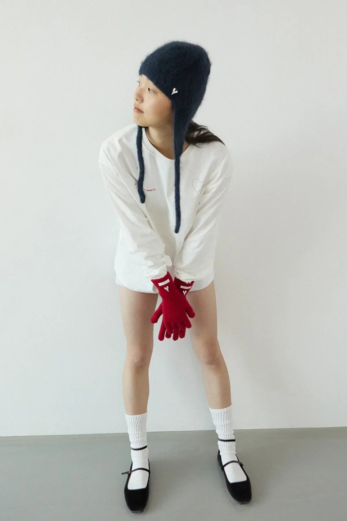 Model wearing red gloves, white long-sleeve shirt, blue knit hat, white socks, and black shoes against plain wall