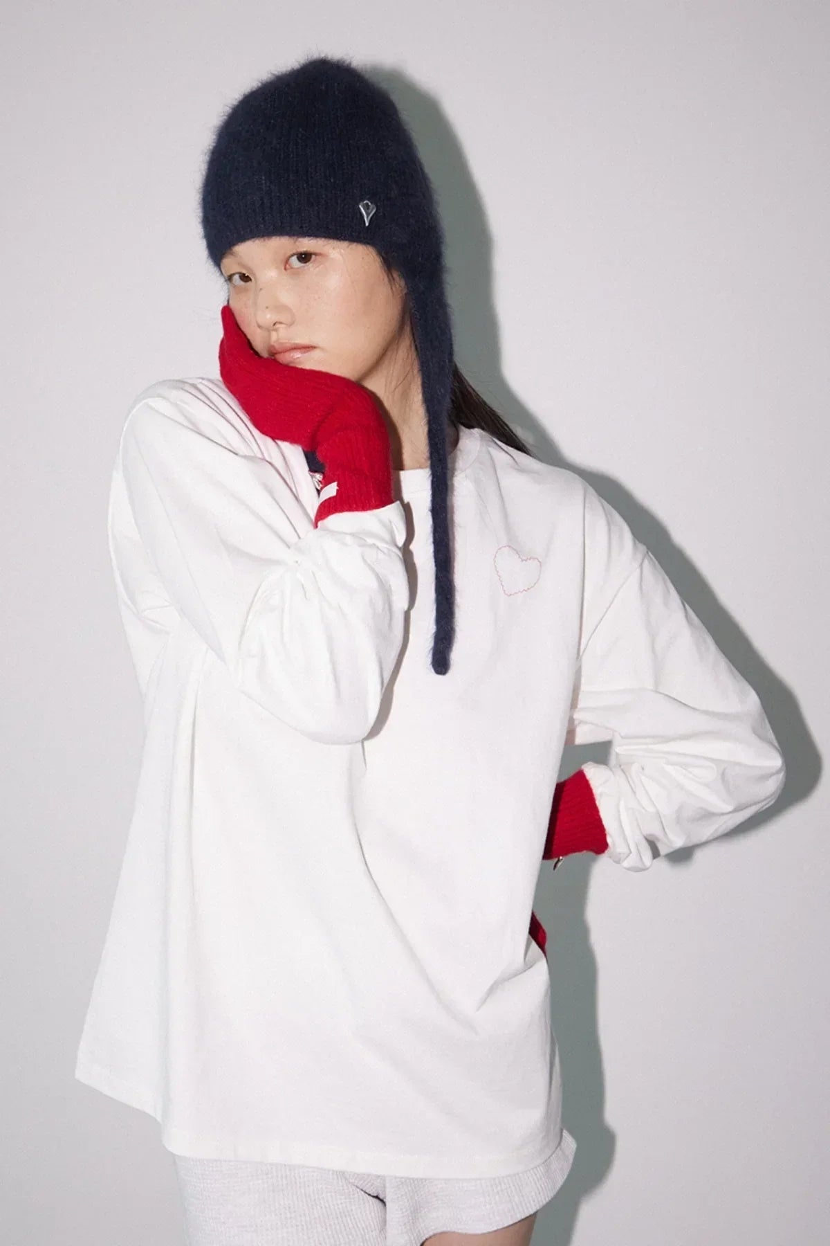 Woman wearing black fuzzy hat, white long sleeve shirt with small heart and red gloves posing against white background