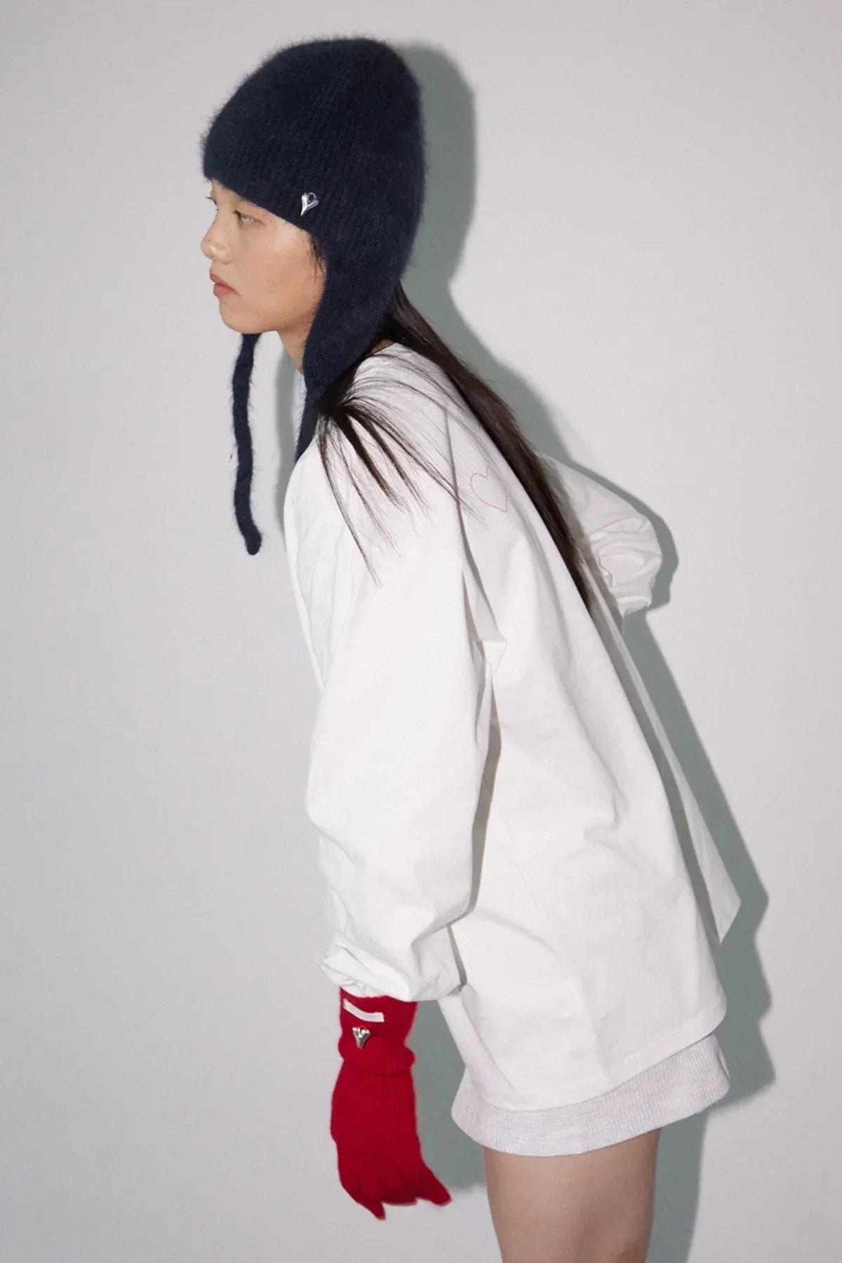 Profile of woman wearing a navy knit hat, white oversized sweatshirt, and red gloves against plain background