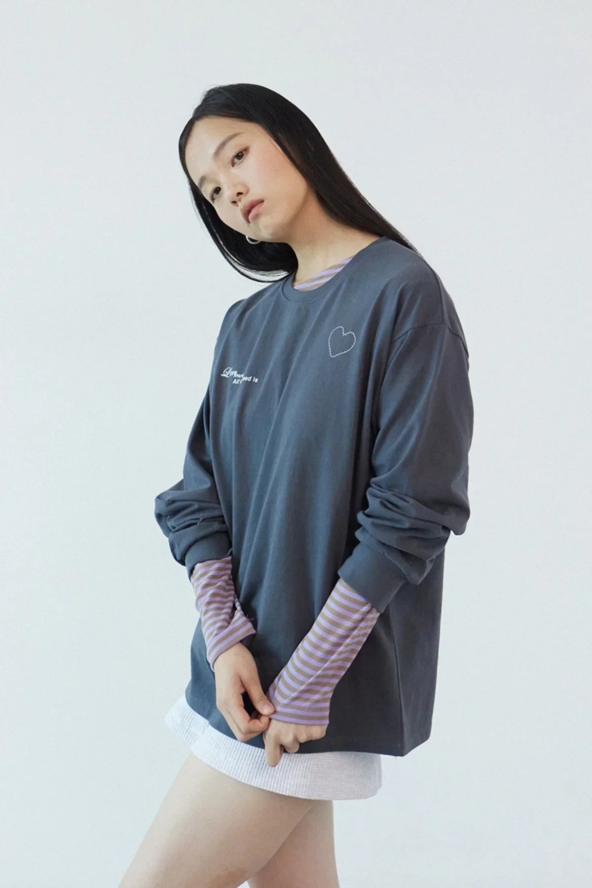 Young woman wearing dark gray long sleeve shirt with small heart and text embroidery, styled with striped inner sleeves and white skirt