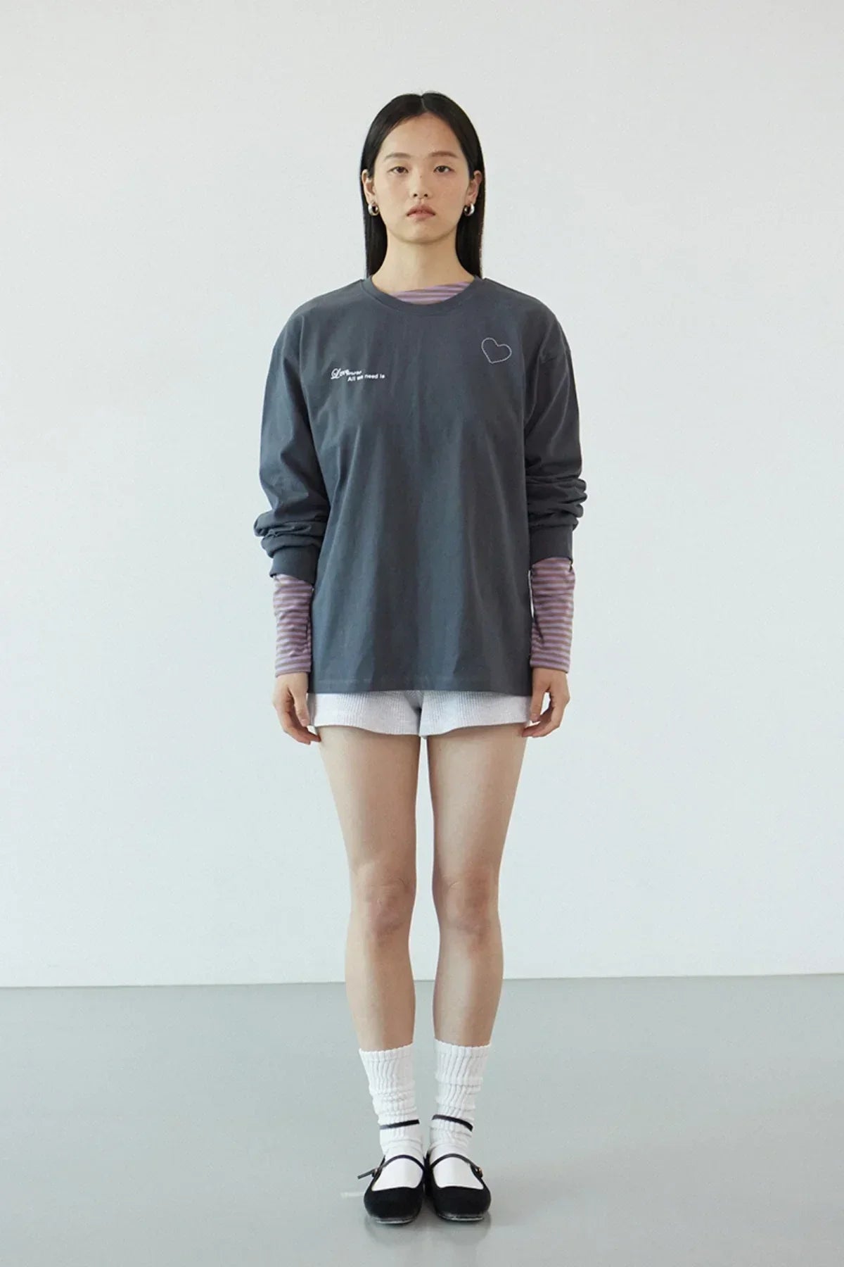 Model wearing dark gray long-sleeve t-shirt with heart and text, striped inner shirt, white shorts, white socks, black shoes