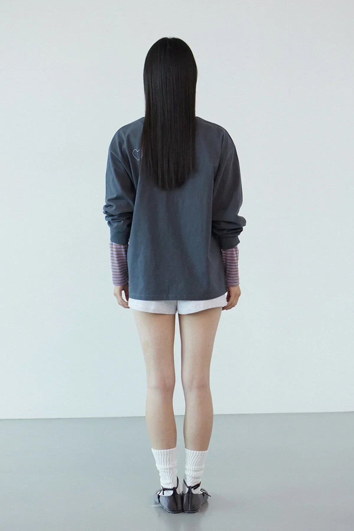 Rear view of woman wearing dark gray long sleeve shirt with heart detail, striped purple sleeves, white shorts, and black shoes
