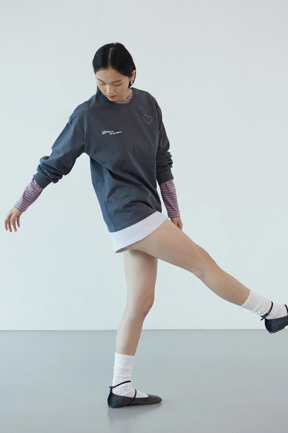 Woman in dark gray long sleeve Love More t-shirt with heart graphic, white shorts, and black flats in studio