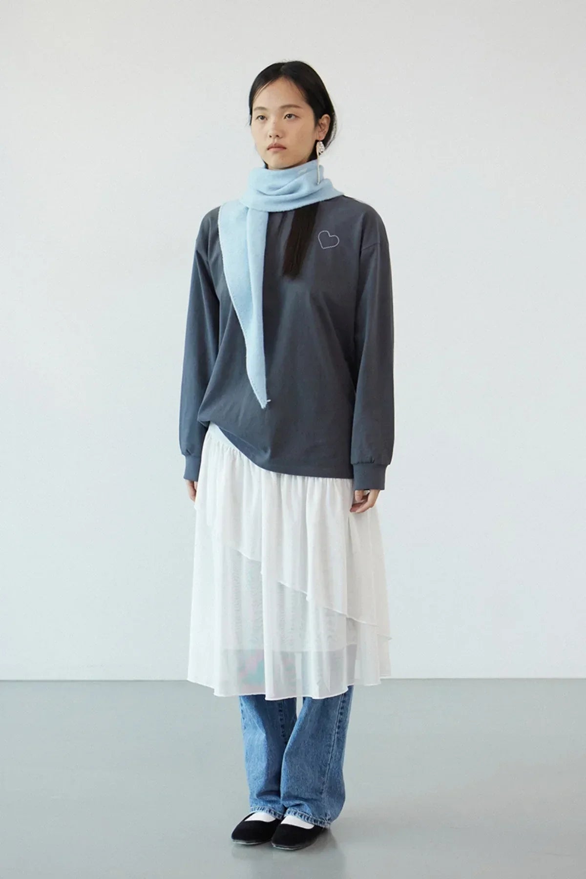 Model wearing dark gray long-sleeve shirt with small heart design, white layered skirt, blue jeans, light blue scarf, and black flats