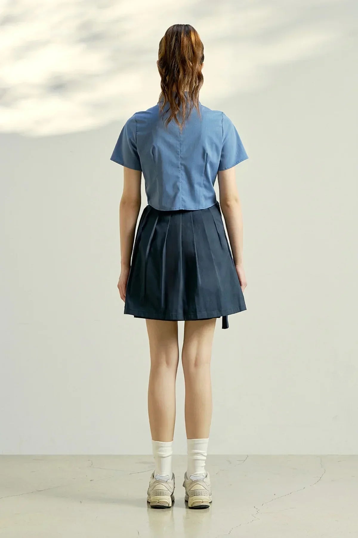Back view of woman wearing indigo blue cropped blouse, navy pleated skirt, white socks, and beige sneakers