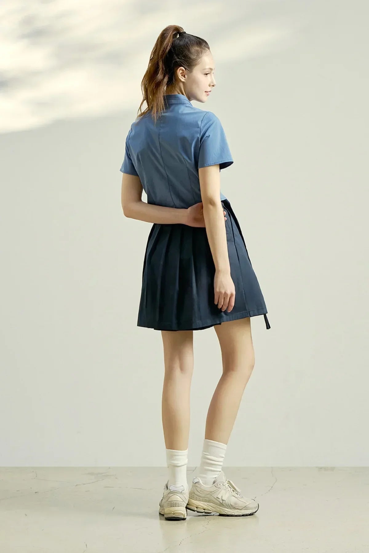Woman wearing indigo blue short-sleeve blouse and pleated skirt, beige sneakers with white socks