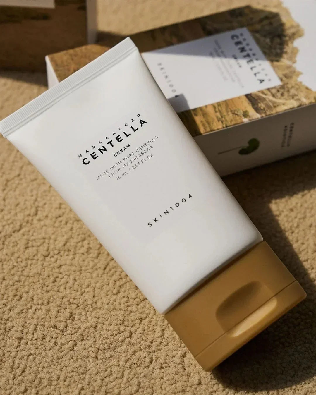 Tube of Madagascar Centella Cream by Skin1004 with brown cap on textured beige surface and product box in background