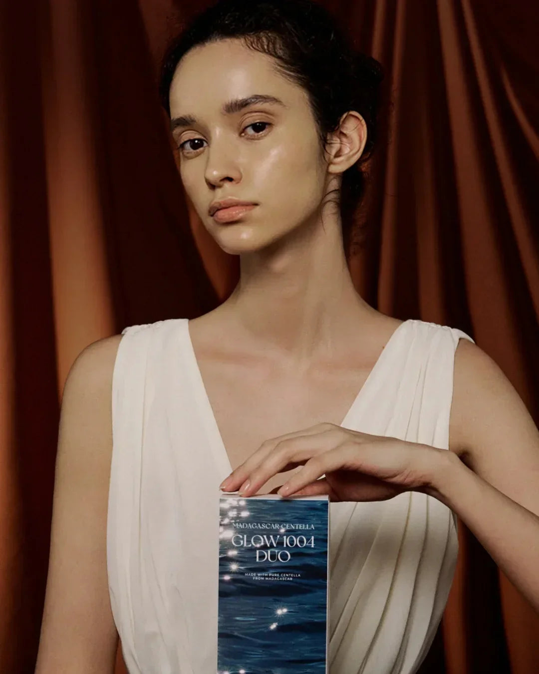 Woman in white dress holding Madagascar Centella Glow 1004 Duo skincare product against brown curtain background