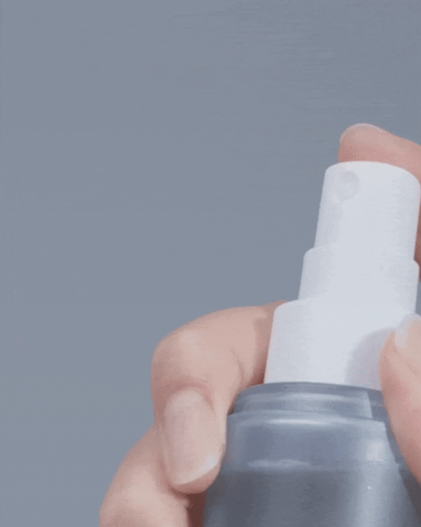 Hand pressing white spray nozzle on gray cosmetic bottle releasing fine moisturizing mist