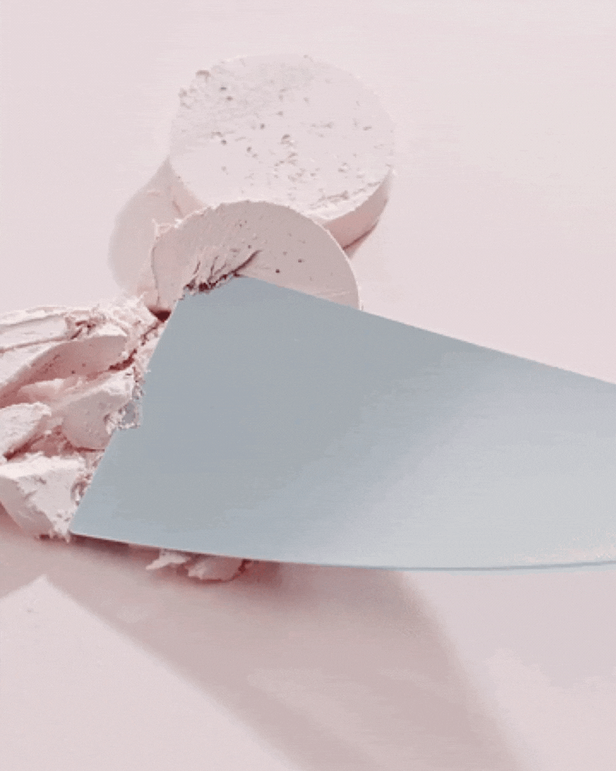 Spatula scraping soft pink clay mask texture from circular clay pieces on pale pink surface