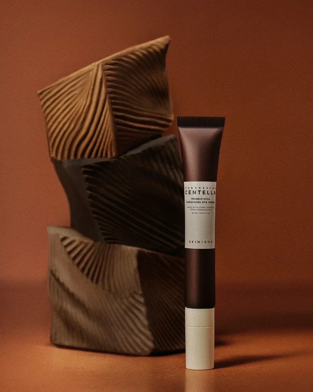 Madagascar Centella Probio-Cica Bakuchiol eye cream tube with sculpted brown textured blocks on brown background