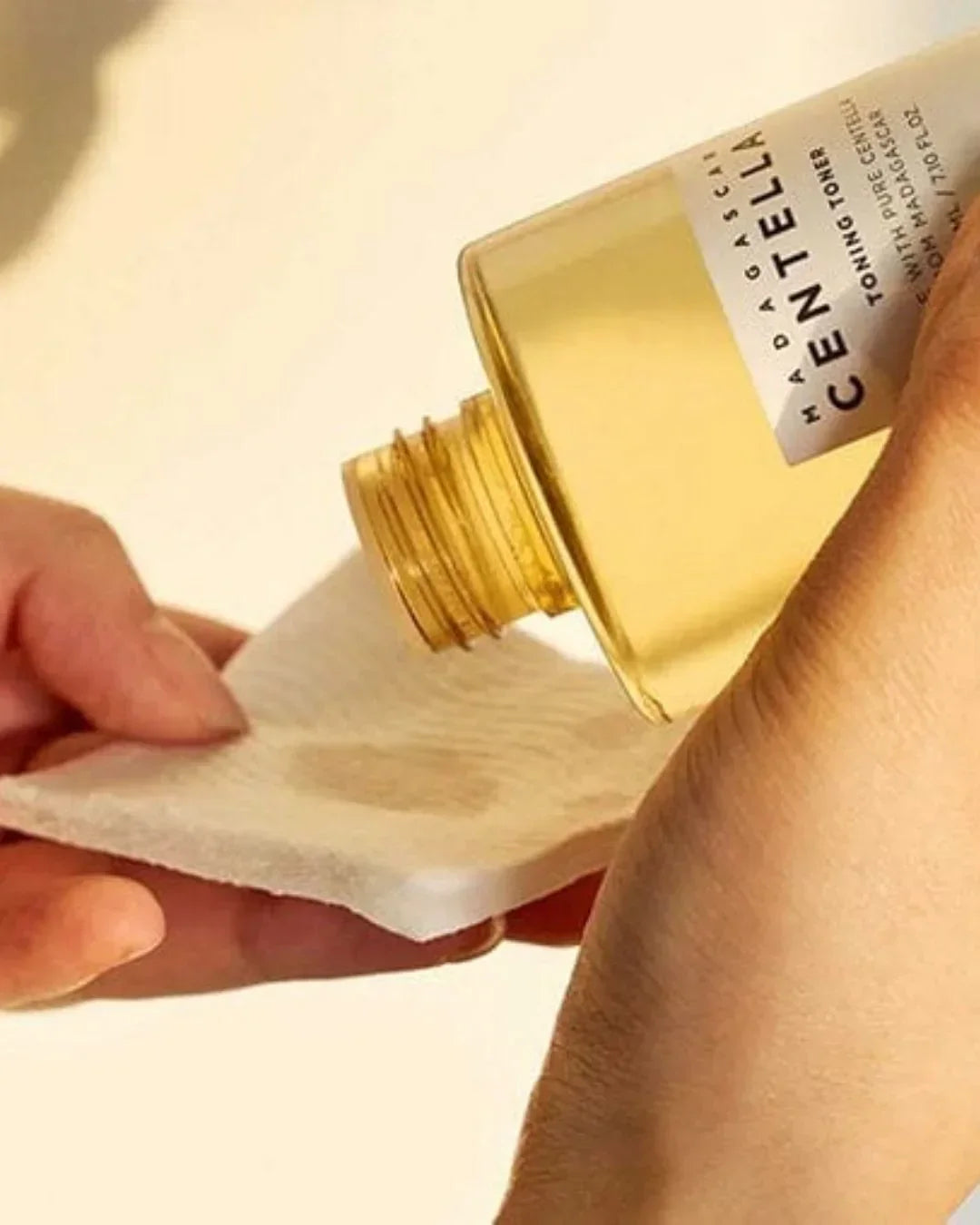 Hand pouring Madagascar Centella toning toner liquid onto white cotton pad against beige background