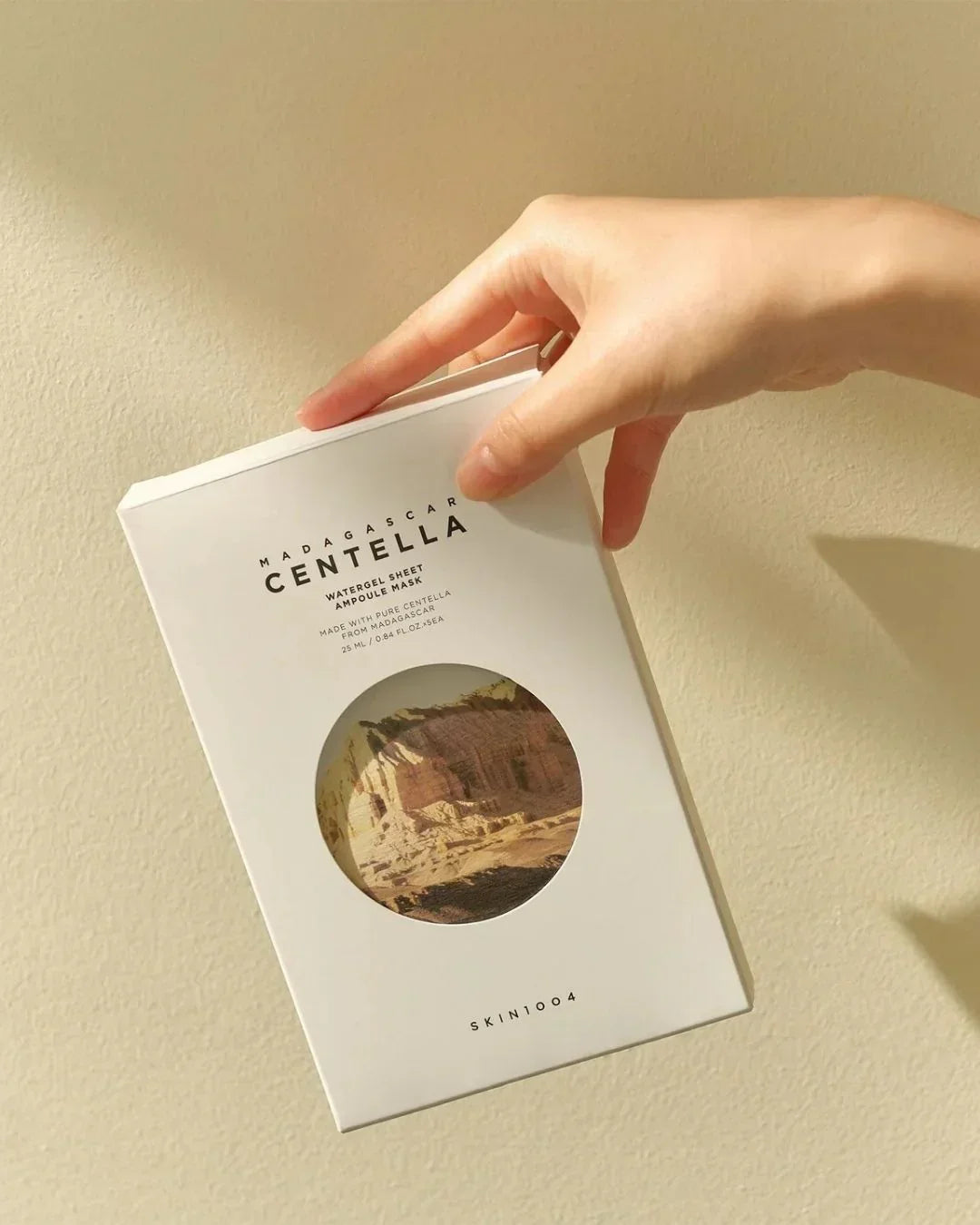 Hand holding SKIN1004 Madagascar Centella Watergel Sheet Ampoule Mask packaging against beige wall