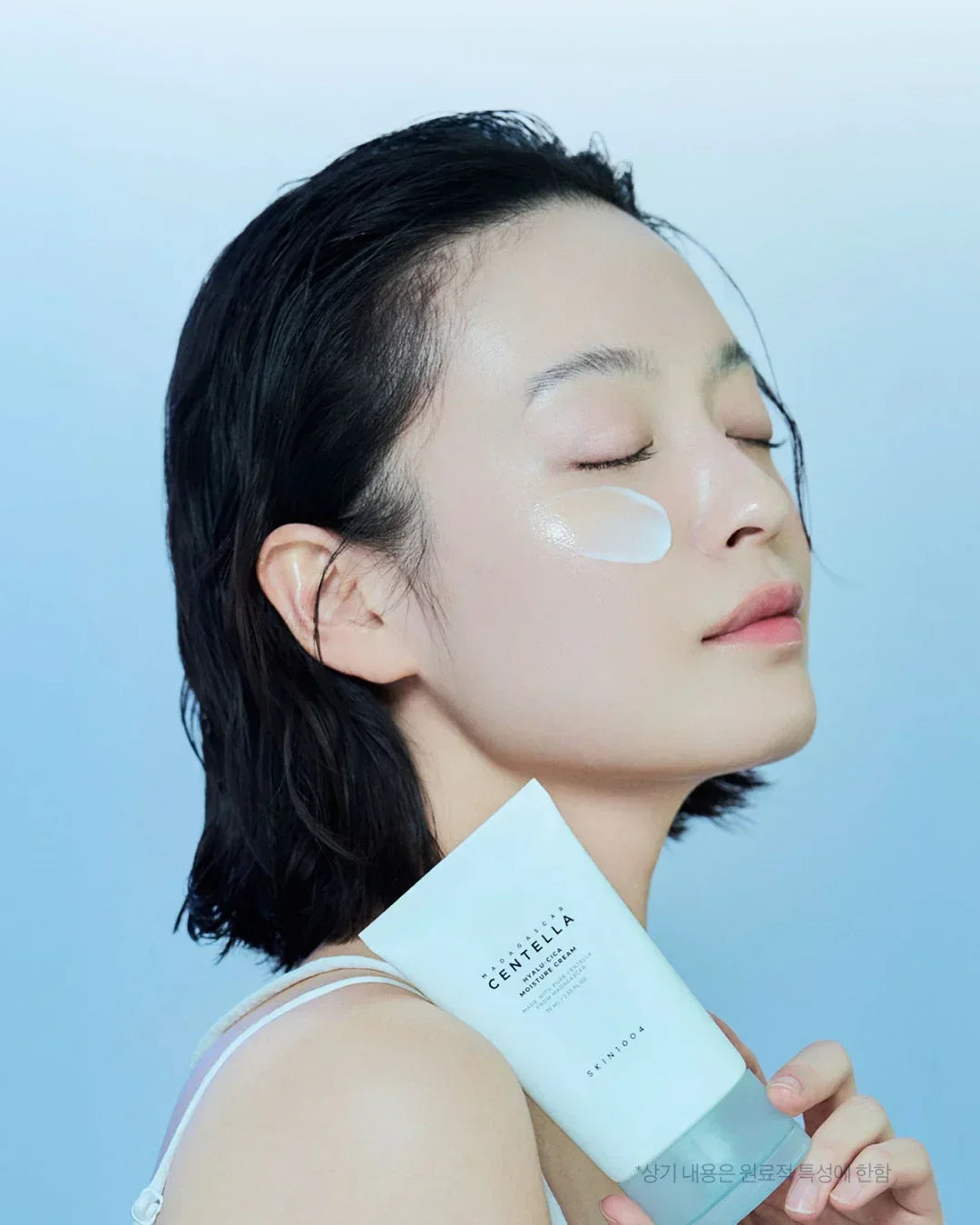 Woman with clear skin applying Madagascar Centella hyaluronic moisture cream on cheek, holding product tube