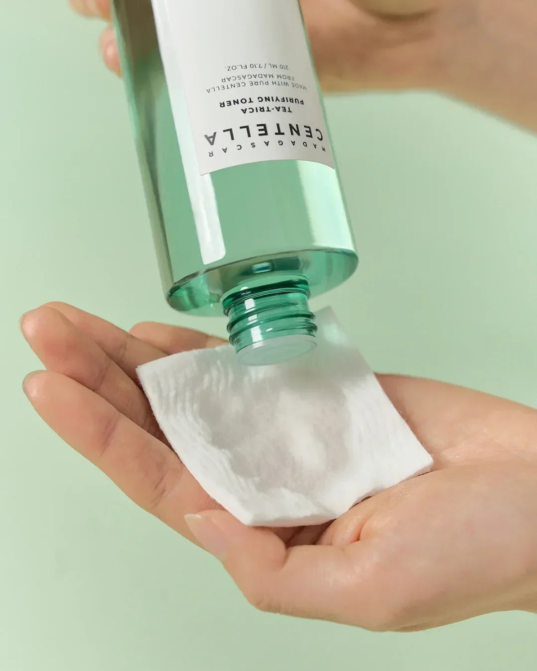 Hand pouring Madagascar Centella Tea-Trica Purifying Toner onto cotton pad with green background