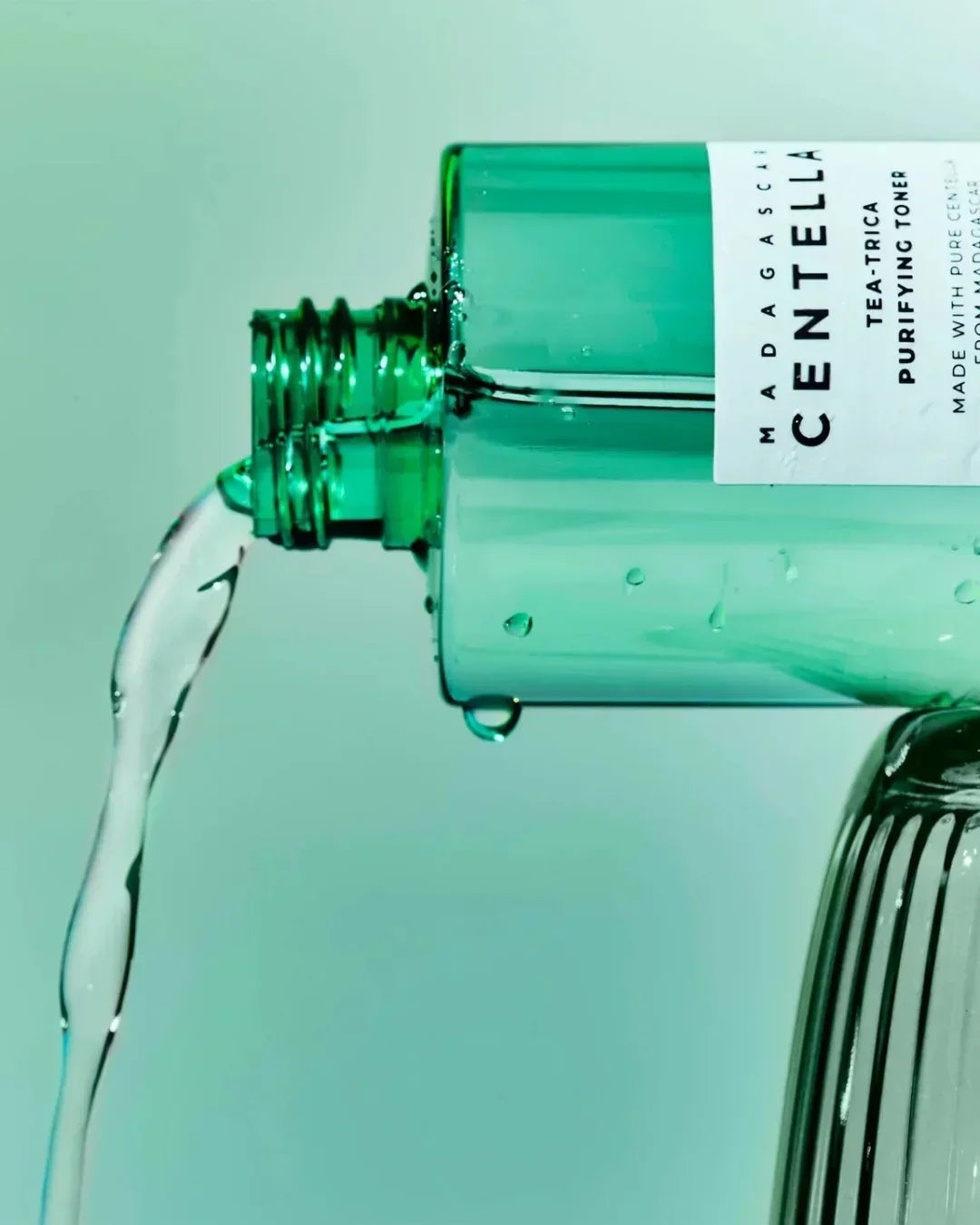 Green bottle of Madagascar Centella Tea-Trica Purifying Toner pouring clear liquid against turquoise background