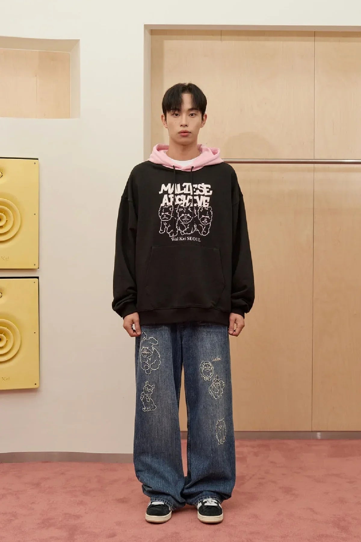 Man wearing black Maltese Archive hoodie with pink hood and blue jeans with dog illustrations in modern indoor setting