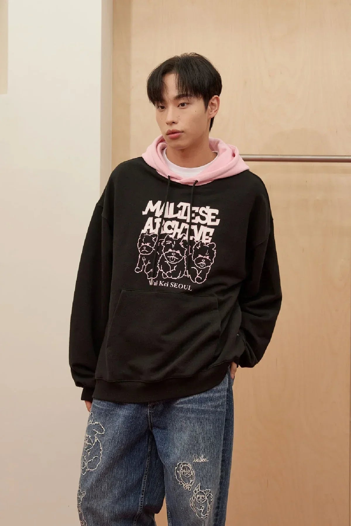 Young man wearing black hoodie with pink dogs graphic and text, paired with embroidered denim jeans