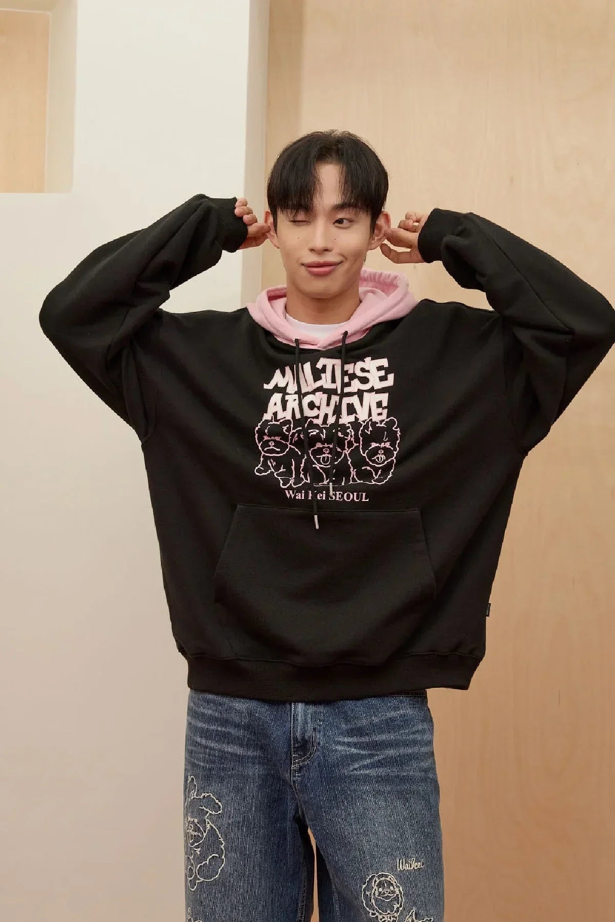 Young man wearing black Maltese Archive hoodie with pink hood and graphic print, paired with embroidered blue jeans
